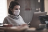 A woman on her computer with a mask on concentrating in a well-lit room.