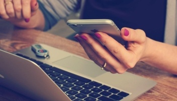 A closeup of a woman with pink nails holding her phone while also on her laptop.